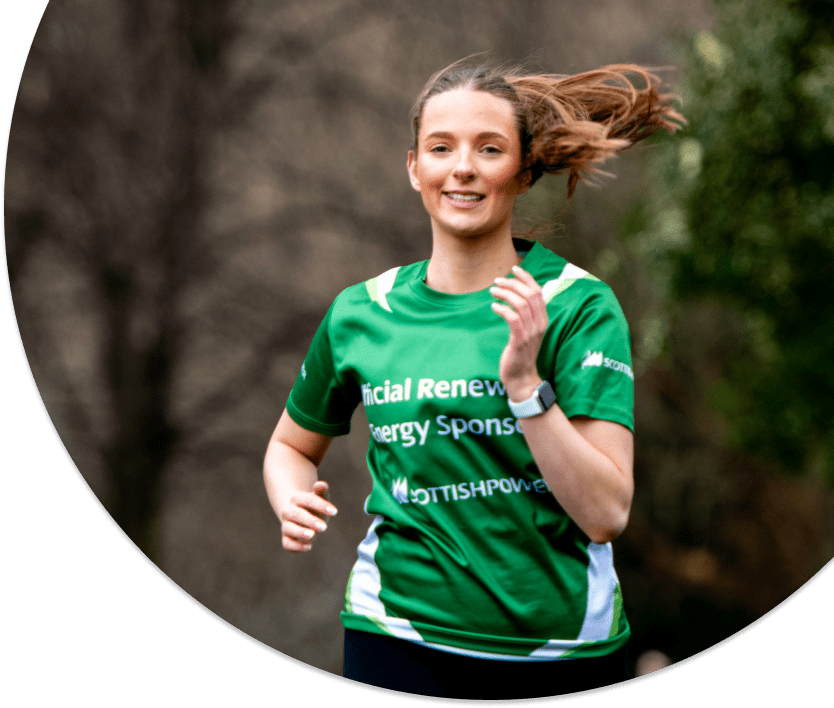 A woman cheerfully running in a park. She is wearing a green Scottish Power Race for Life jersey.